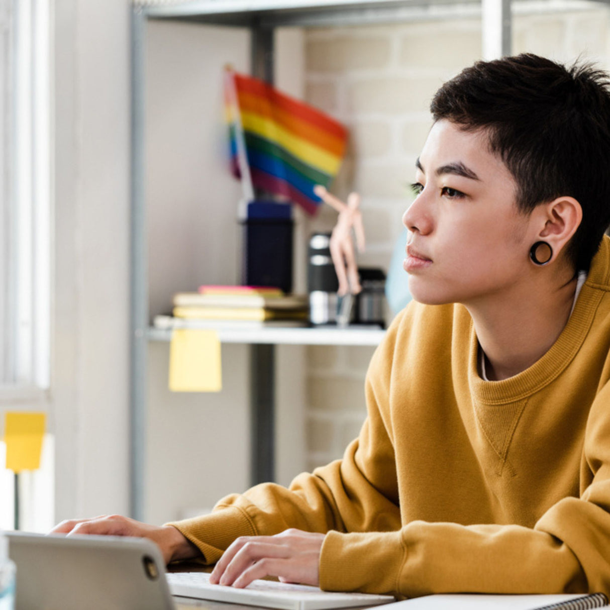 Person wearing a yellow sweater sitting at a desk with a laptop, rainbow flag in the background